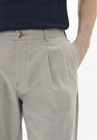 Pantalones de vestir beige claro con una textura suave, con pliegues en la parte frontal, bolsillos laterales y un cierre de botón único en la cintura.