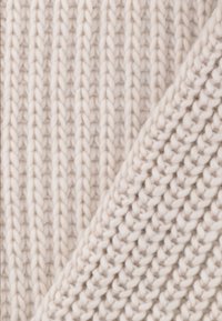 Gruba, kremowa dzianina z wyraźnym pionowym prążkowaniem, o gładkiej teksturze i złożonym brzegu, ukazującym inny wzór dzianiny.