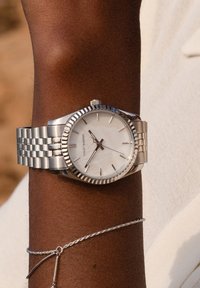 Silberner Edelstahluhren mit einem runden Zifferblatt aus weißem Perlmutt, silbernen Stundenmarkierungen und einem strukturierten Gehäuse. Armband aus Metallgliedern.