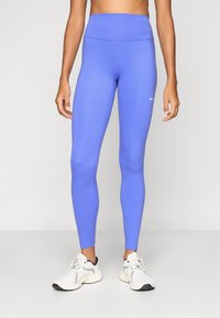 Κολάν Nike σε έντονο μπλε χρώμα, με ψηλή μέση, λεία υφή και μικρό λευκό λογότυπο στο αριστερό κάτω μέρος του ποδιού. Συνδυάζεται με λευκά αθλητικά παπούτσια.