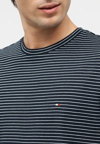 Κάτω μέρος του προσώπου και ο λαιμός ενός άνδρα, που φοράει μπλε σκούρο πουκάμισο με λεπτές οριζόντιες λευκές ρίγες και ένα μικρό ορθογώνιο κόκκινο και άσπρο λογότυπο στο στήθος.