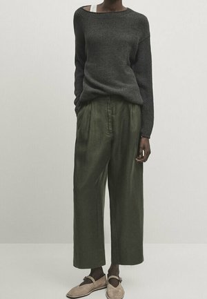 Persoon die een donkergrijze gebreide trui draagt, losse olijfgroene broek en beige platte schoenen, staand tegen een effen lichte achtergrond.