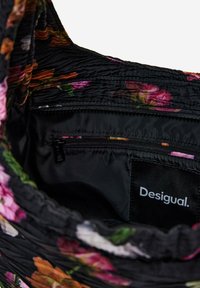 Sisäpuoli mustasta kukkakuosisesta laukusta, jossa on vaaleanpunaisia ja oransseja kukkia. Sisältää vetoketjullisen taskun, tekstuurillista kangasta ja tarran, jossa lukee "Desigual."