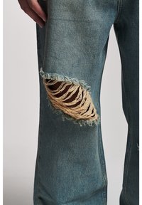 Modré džíny z opotřebovaného denimu s velkou vytahanou dírou na koleni, která odhaluje světlejší vnitřní látku. Mají volný střih a texturovaný povrch.