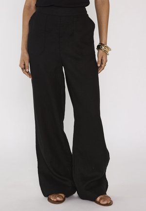 Personne portant un pantalon noir ample, un haut noir, des sandales marron, des bracelets dorés et des bagues, debout devant un fond blanc uni.