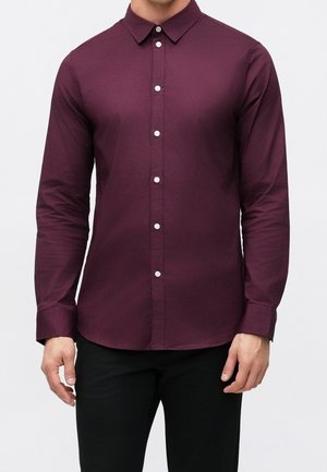 Mann trägt ein eng anliegendes, langärmeliges Hemd in dunklem Burgund mit weißen Knöpfen und schwarze Hose, steht vor einem einfarbigen Hintergrund.