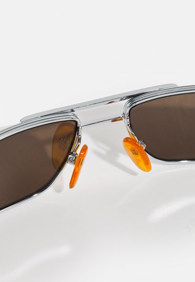 Lunettes de soleil avec une monture chromée, des verres bruns et des plaquettes de nez orange ; elles présentent un design à double verre avec une forme géométrique élégante.