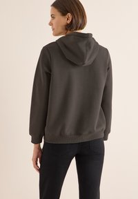 Felpa con cappuccio di colore verde scuro, realizzata in tessuto misto cotone. Presenta un cappuccio con cordino e polsini a coste. Indossata con jeans neri, vista da dietro.
