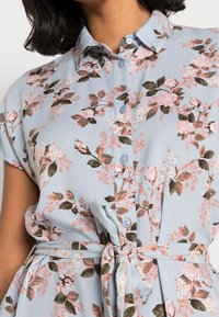 Donna che indossa un vestito floreale azzurro chiaro con maniche corte, bottoni frontali, colletto e cintura di stoffa legata in vita.