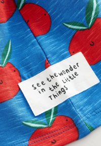 Tissu bleu avec des motifs rouge orangé vif et des feuilles vertes. Une étiquette blanche avec du texte noir indique : « Voir la merveille dans les petites choses. »