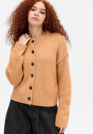Junge Frau mit lockigem roten Haar, die einen geknöpften beigen Wollpullover und schwarze, weit geschnittene Nadelstreifen-Hosen vor weißem Hintergrund trägt.