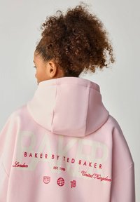 Rozā kapuce ar lielu "BAKER" tekstu bēšā krāsā, sarkaniem logo detaļām un kapuci. Mīksts audums ar brīvu piegriezumu un teksturētām akcentiem.