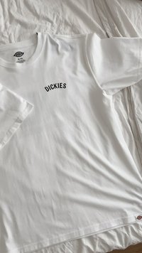 Hvit t-skjorte med en svart "DICKIES" logo i et buet skrifttype på brystet. Laget av myk bomull, med en avslappet passform og korte ermer.