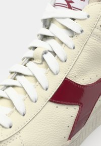 Cream leather sneaker with a glossy red side logo, white laces, and textured surface. Features striped ear and perforations along the upper.