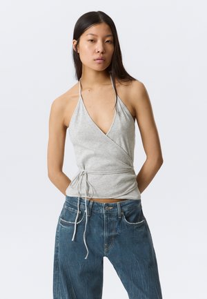 Mujer joven con cabello largo y oscuro que lleva un top gris halter anudado a la cintura y vaqueros azules sueltos sobre un fondo liso.