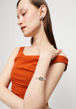 Boucles d'oreilles en argent et un bracelet avec un détail circulaire. La chemise est orange, avec un col en U, et il y a deux lanières blanches sur le bracelet.