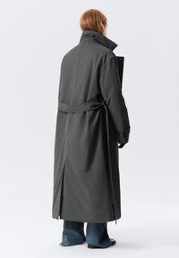Lungo cappotto grigio con colletto alto, vita cintura e fessure laterali. Indossato sopra jeans blu a gamba larga. Tessuto liscio e preciso.