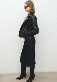 Czarna skórzana kurtka motocyklowa, ciemna spódnica z denimu oraz czarne kozaki za kolano. Długi bob i przyciemniane, duże okulary przeciwsłoneczne dopełniają stylizację.