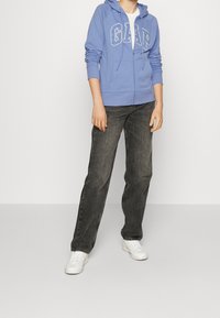 Haut à capuche zippé bleu clair avec le logo "GAP", associé à un jean gris foncé et des baskets blanches. Tissu lisse, coupe décontractée et design simple.