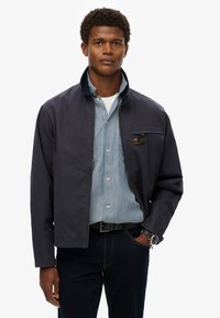 Marineblå bomuldsjakke med ståkrave, lynlås og brystlomme. Båret over en stribet skjorte med knapper og mørke jeans.