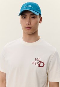 Krēmkrāsas kokvilnas T-krekls ar tumši sarkanu "LD" logotipu uz kreisā krūšu rajona, kopā ar tirkīza samta cepuri ar baltu izšūtu tekstu.