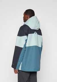 Casaco à prova d'água com um design de blocos de cores em azul marinho, azul claro e teal. Apresenta capuz e punhos ajustáveis. O material parece texturizado.