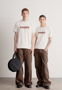 Dwie modelki noszą białe bawełniane koszulki z czerwonym logo „tommy jeans”, połączone z brązowymi szerokimi spodniami. Jedna z nich trzyma czarną okrągłą torebkę.