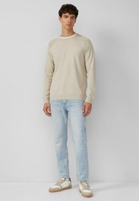 Hellbeige gestrickter Pullover mit langen Ärmeln, rundem Ausschnitt und strukturiertem Stoff. Kombiniert mit hellblauen Jeans und weißen Sneakers mit beigen Akzenten.