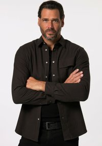 Camicia marrone con bottoni in tessuto morbido, dotata di tasca sul petto, bottoni argentati e colletto classico, indossata sopra una maglietta nera.