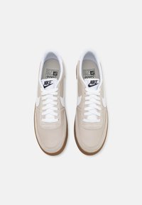 Beiga lädersneakers med vita detaljer och gummisula. Har vita skosnören och en märkeslogotyp på tungan. Ren, minimalistisk design.