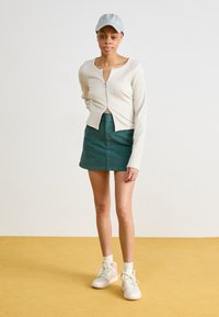 Κρεμ καπιτονέ κοντό πουλόβερ με φερμουάρ μπροστά, συνδυασμένο με μια τιρκουάζ μίνι φούστα και λευκά αθλητικά παπούτσια με ροζ λεπτομέρειες. Απαλό μπλε καπέλο.