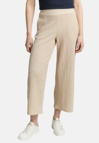 Pantalons larges beiges, coupés, en tissutexturé avec un motif en relief, associés à des baskets blanches. Ceinture élastique pour le confort.