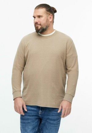 Homme avec barbe et chignon, portant une chemise beige à manches longues et un jean bleu, debout et regardant vers la gauche sur fond blanc.