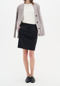 Cardigan en tricot gris, top blanc et jupe noire mi-longue. Cardigan avec texture côtelée et boutons ; jupe simple et structurée ; chaussures noires à enfiler.