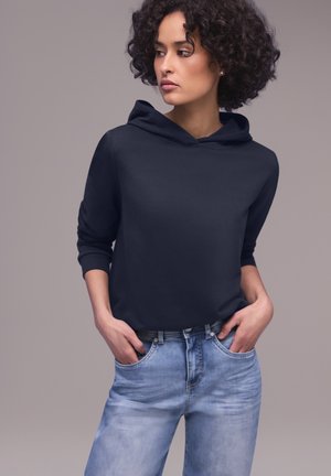 Donna con capelli ricci che indossa una felpa con cappuccio blu scuro e jeans azzurri, con le mani nelle tasche, che guarda di lato su uno sfondo semplice.