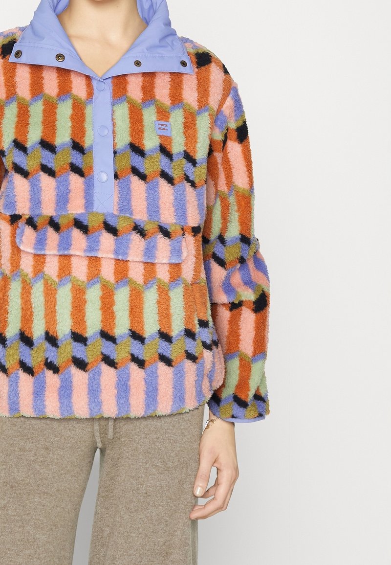Pull polaire multicolore avec motif géométrique, patte de boutonnage et col lavande, porté avec un pantalon beige sur un fond clair.