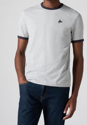 Homme portant un t-shirt gris clair à manches courtes avec bordure foncée et petit logo noir sur la poitrine, associé à un jean bleu foncé, main dans la poche.