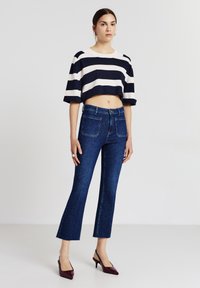 Top a righe corto in maglia di colore blu navy e bianco, abbinato a jeans in denim scuri a palazzo con tasche frontali e posteriori; il modello indossa tacchi alti.