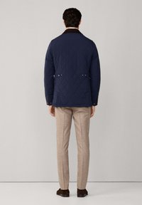 Gequiltete navyblaue Jacke mit einem braunen Cordkragen, Druckknöpfen und Seitentaschen, kombiniert mit beigen Hosen und braunen Schuhen.