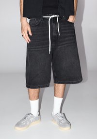 Shorts en denim noir avec une coupe ample, dotés d'une taille élastique avec cordon blanc. Associés à des baskets grises et des chaussettes blanches aux chevilles.