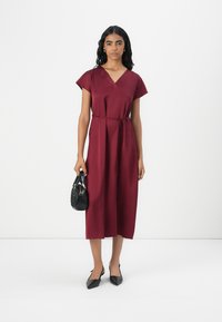 Robe bordeaux avec des manches courtes, décolleté en V, ceinture à nouer à la taille et longueur midi. Portée avec des escarpins noirs à bout pointu et un petit sac à main noir.