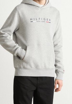 Osoba ubrana w jasnoszarą bluzę z kapturem z napisem i logo "HILFIGER NEW YORK", w czarnych spodniach, stojąca na jednolitym tle.