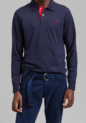 Marineblaues Polohemd mit langen Ärmeln und kontrastierendem roten Kragen, besticktem Logo auf der Brust, kombiniert mit dunkler Hose und blau gestreiftem Gürtel.