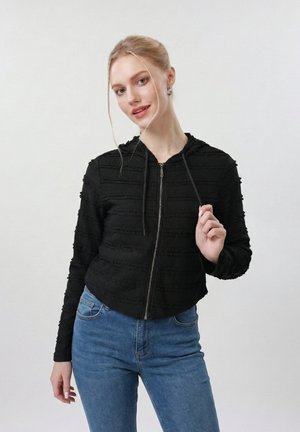 Fiatal nő világosbarna hajjal, alacsonyan összefogott kontyban, fekete, texturált cipzáros kapucnis pulóvert és kék farmert visel, egyik kapucnis zsinórt tartja a kezében.