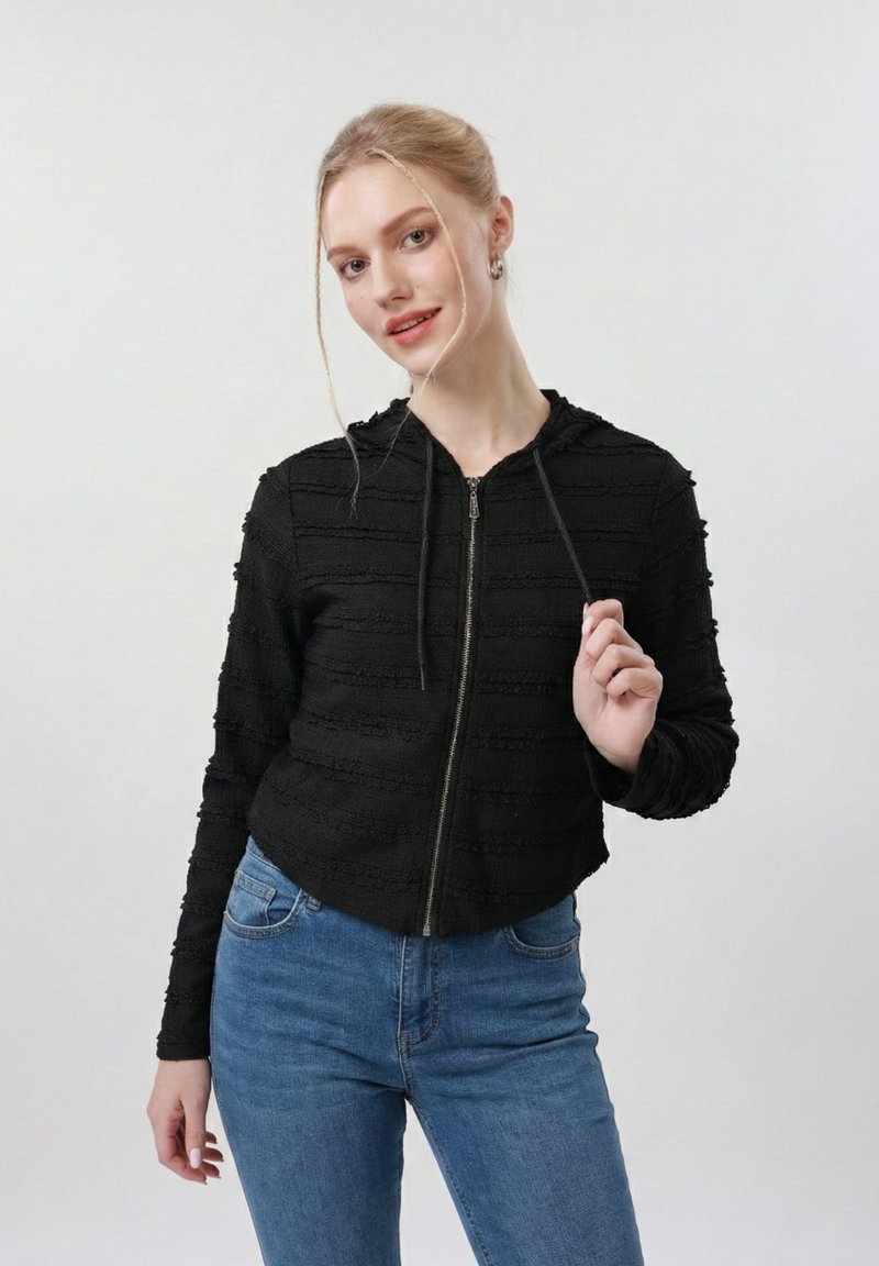 Νεαρή γυναίκα με ξανθά μαλλιά σε χαμηλό κότσο φοράει μια μαύρη υφασμάτινη ζακέτα με φερμουάρ και μπλε τζιν, κρατώντας μία από τις κορδόνια της ζακέτας.