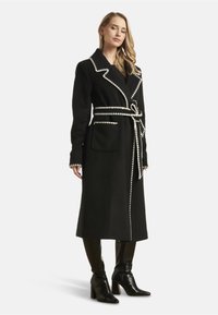 Black long coat with contrasting white trim, wide lapels, and a belt. Features two front pockets and a structured fit. Worn with knee-high boots.