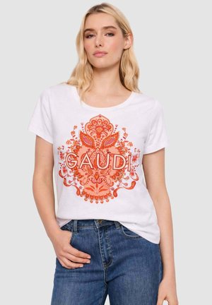 Donna bionda che indossa una maglietta bianca con motivo floreale arancione e la scritta "GAUDI", abbinata a jeans blu, in piedi contro uno sfondo chiaro.