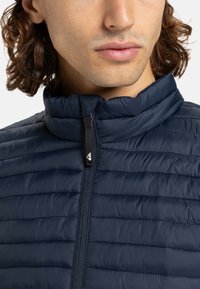 Mann mit schulterlangem Haar, der eine navyblaue gesteppte Jacke mit Stehkragen und Reißverschluss trägt, sichtbar von Kinn bis zu den Schultern.