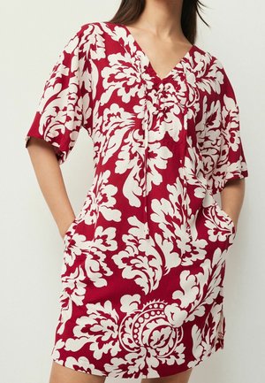 Femme portant une robe rouge avec de grands motifs floraux blancs, manches courtes, col en V avec liens, et les mains dans les poches latérales.