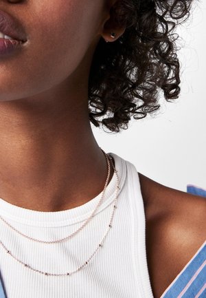 Primer plano de una persona que lleva dos delicados collares de oro y una blusa blanca de canalé debajo de una prenda a rayas azul y rosa.